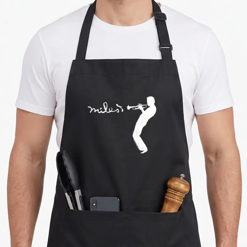 Tablier de cuisine noir avec silhouette blanche d'un musicien jouant du trompette et l'inscription miles en écriture cursive à côté.