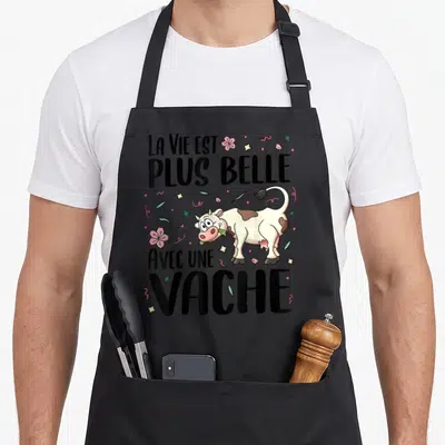 Tablier de cuisine noir illustré d'une vache rieuse mâchant de l'herbe entourée de fleurs roses et d'étoiles multicolores.
