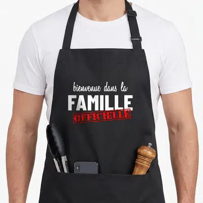 Tablier de cuisine noir avec inscription bienvenue dans la famille « officielle » en blanc et rouge.