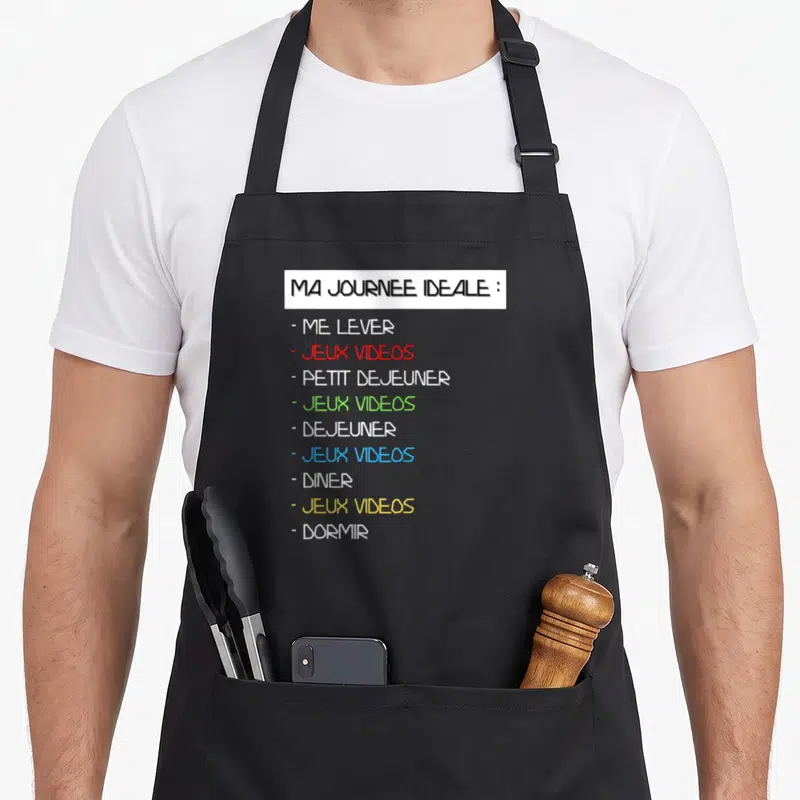 Tablier de cuisine noir avec liste imprimée Ma journée idéale alternant repas et jeux vidéos en couleurs vives sur fond blanc.