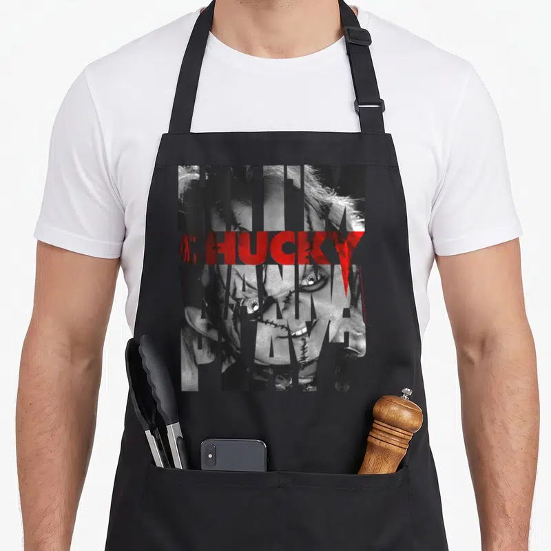 Tablier de cuisine noir avec l'illustration Hi I'm Chucky Wanna Play en texte et visage en noir et blanc.