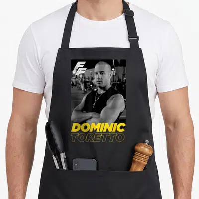 Tablier de cuisine noir avec portrait de Dominic Toretto, logo F&F et nom en lettres jaunes audacieuses sur fond urbain flou.