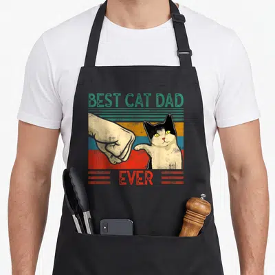 Tablier de cuisine noir avec illustration vintage d'un chat tuxedo et d'une main effectuant un poing contre poing sur fond coloré.
