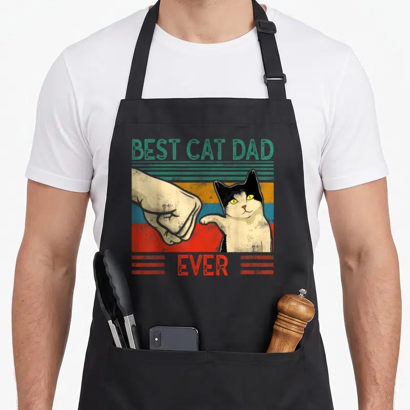 Tablier de cuisine noir avec illustration vintage d'un chat tuxedo et d'une main effectuant un poing contre poing sur fond coloré.