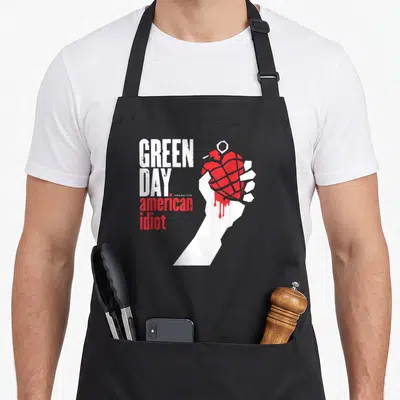 Tablier de cuisine noir avec illustration d'une main blanche tenant une grenade rouge et le texte Green Day American Idiot en blanc et rouge.