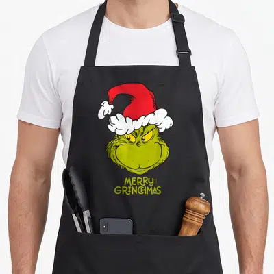 Tablier de cuisine noir avec le visage vert du Grinch coiffé d'un bonnet rouge et blanc.