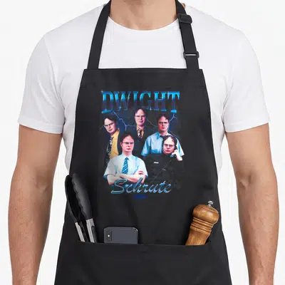 Tablier de cuisine noir avec le nom Dwight Schrute en lettres bleues et cinq visages du personnage entourés d'éclairs.