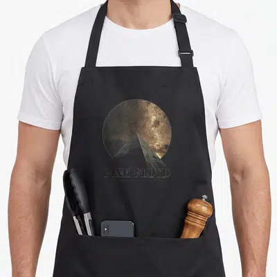 Tablier de cuisine noir avec une pyramide blanche devant un disque galactique doré et le nom du groupe imprimé en dessous.
