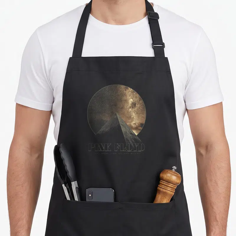 Tablier de cuisine noir avec une pyramide blanche devant un disque galactique doré et le nom du groupe imprimé en dessous.