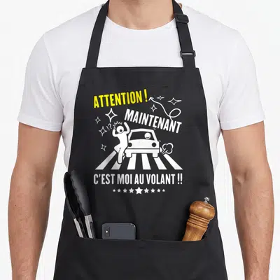 Tablier de cuisine noir avec une illustration blanche d'une voiture et du texte jaune et blanc humoristique.