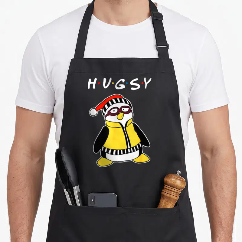 Tablier de cuisine noir avec un pingouin nommé Hugsy portant un chapeau rayé et des lunettes violettes.