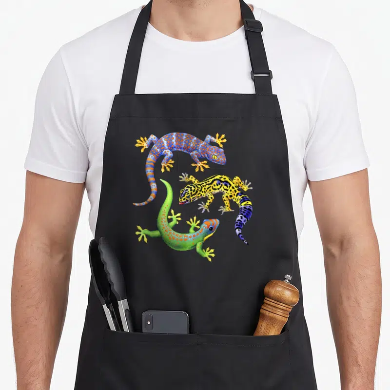 Tablier de cuisine noir avec trois lézards aux couleurs vives et motifs géométriques distincts.
