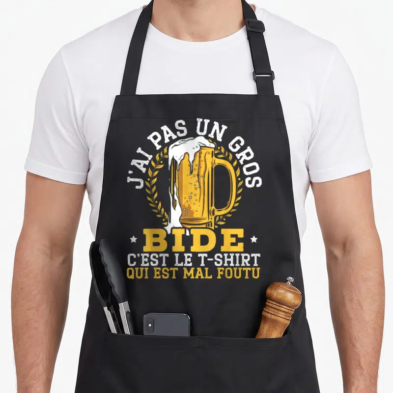 Tablier de cuisine noir avec illustration de grande bière déversée entourée de texte humoristique blanc et jaune.