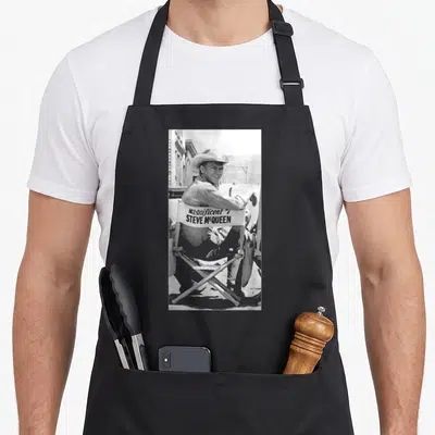 Tablier de cuisine noir avec une illustration en noir et blanc de Steve McQueen assis sur une chaise de tournage.