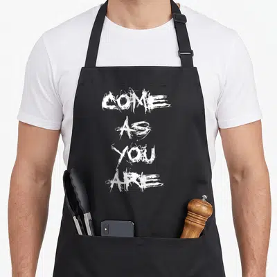 Tablier de cuisine noir avec inscription « Come As You Are » en lettres blanches style graffiti vertical.