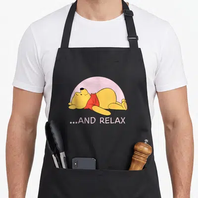 Tablier de cuisine noir avec Winnie l'Ourson jaune en tenue rouge allongé sur fond demi-cercle rose et texte...AND RELAX.