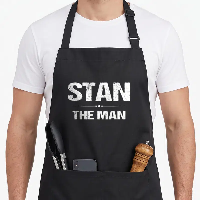Tablier de cuisine noir avec l'inscription blanche « STAN THE MAN » en lettres capitales au centre du vêtement.