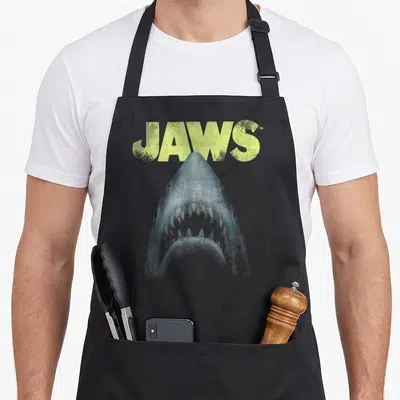 Tablier de cuisine noir avec illustration d'un grand requin blanc et le mot Jaws en lettres jaunes au-dessus.