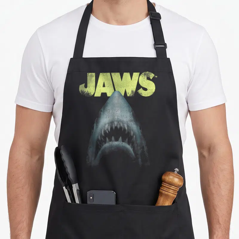 Tablier de cuisine noir avec illustration d'un grand requin blanc et le mot Jaws en lettres jaunes au-dessus.