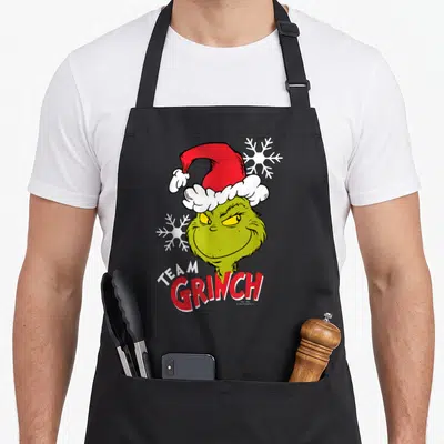 Tablier de cuisine noir avec illustration du Team Grinch portant un bonnet rouge et des flocons blancs.