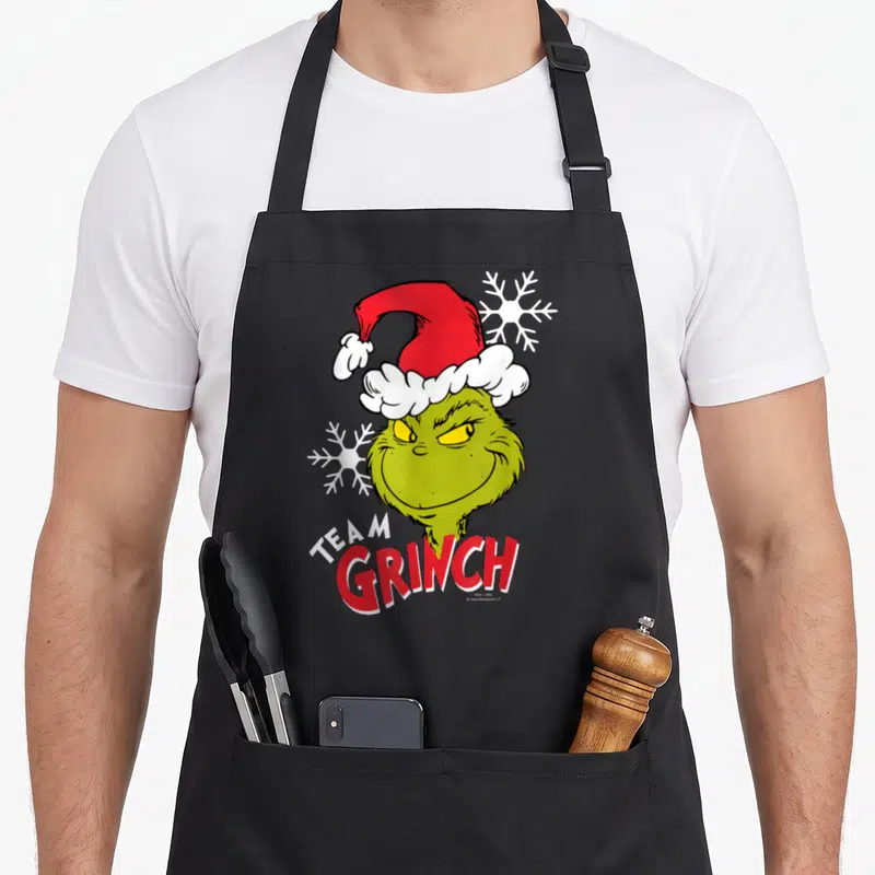 Tablier de cuisine noir avec illustration du Team Grinch portant un bonnet rouge et des flocons blancs.