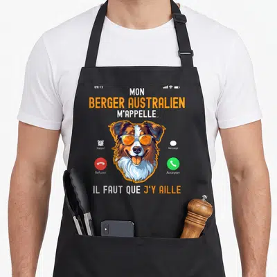 Tablier de cuisine noir avec illustration d'un Berger Australien portant des lunettes orange et du texte humoristique sur fond gris.