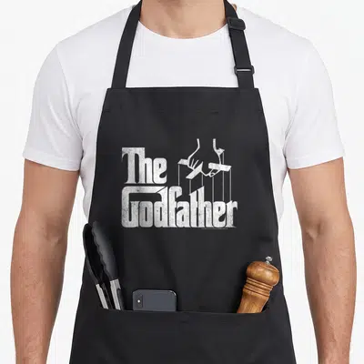 Tablier de cuisine noir avec le logo The Godfather en blanc texturé et une main de marionnettiste stylisée au-dessus des lettres.