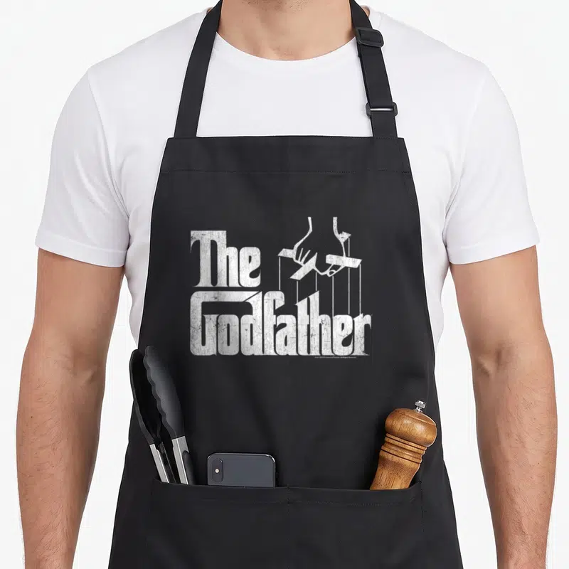 Tablier de cuisine noir avec le logo The Godfather en blanc texturé et une main de marionnettiste stylisée au-dessus des lettres.