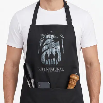 Tablier de cuisine noir avec trois silhouettes humaines devant des arbres et le texte supernatural join the hunt.