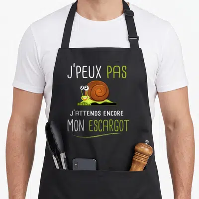 Tablier de cuisine noir avec un escargot vert souriant et le texte J'peux pas j'attends mon escargot imprimé en blanc et vert.