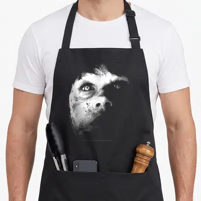 Tablier de cuisine noir avec illustration monochrome du visage de Regan le démon, style grainé et terrifiant.
