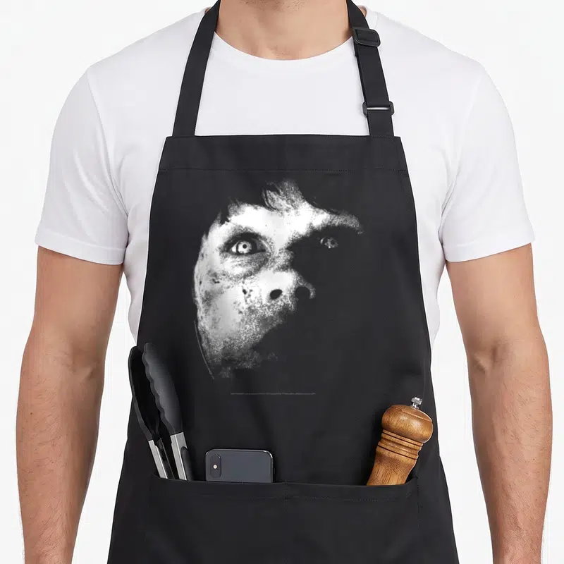 Tablier de cuisine noir avec illustration monochrome du visage de Regan le démon, style grainé et terrifiant.