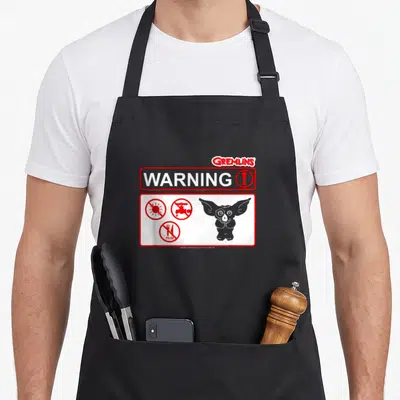 Tablier de cuisine noir avec panneau d'avertissement Gremlins et pictogrammes rouges interdits.