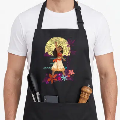 Tablier de cuisine noir avec Vaiana levant la pierre verte devant un motif circulaire jaune et des fleurs tropicales violettes et orange.