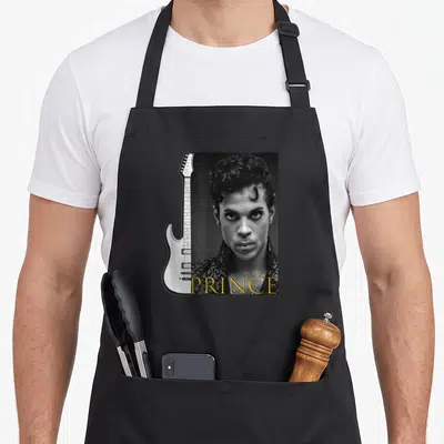 Tablier de cuisine noir avec portrait Prince, guitare électrique blanche et inscription dorée PRINCE en bas à droite.