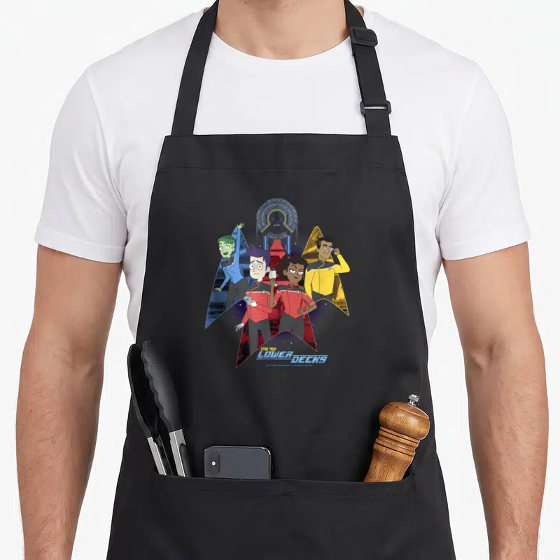 Tablier de cuisine noir avec quatre personnages animés en uniformes colorés devant un insigne spatial stylisé et un dôme technique.