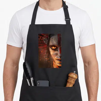 Tablier de cuisine noir avec portrait d'un personnage aux yeux dorés et visages peints en rouge et blanc.