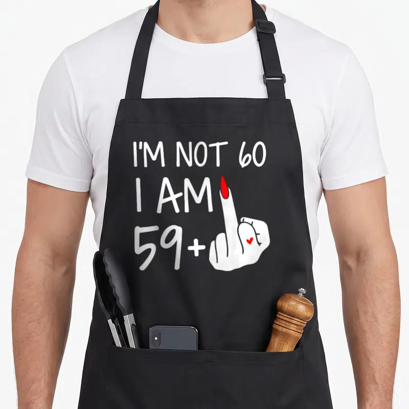 Tablier de cuisine noir avec texte blanc Im Not 60 I Am 59 Plus 1 et illustration de main avec ongle rouge et petit cœur.