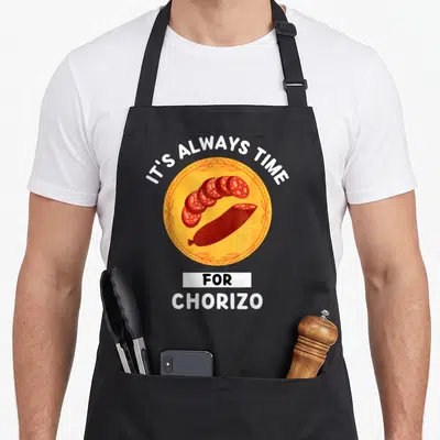 Tablier de cuisine noir avec une illustration circulaire orange représentant un chorizo et le texte It's Always Time For Manger Chorizo.