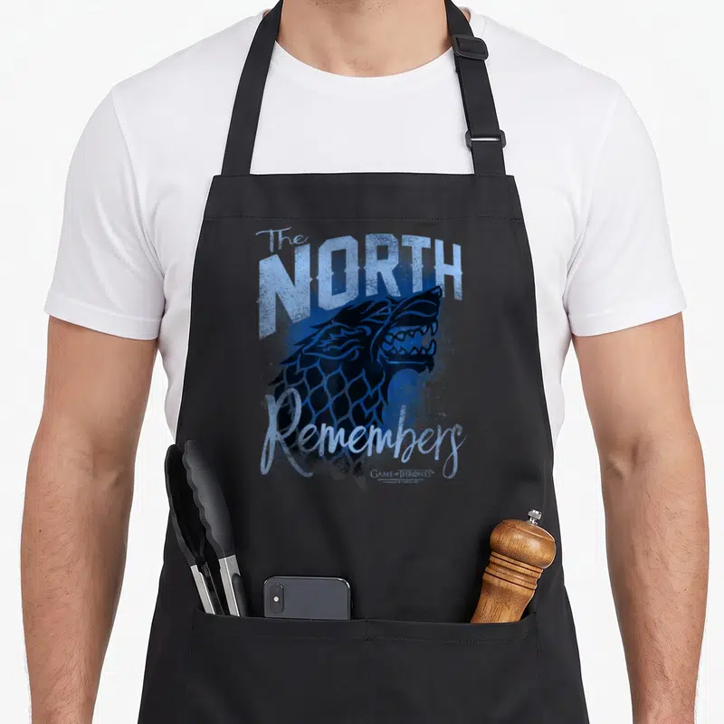 Tablier de cuisine noir avec un loup hurlant bleu et le texte The North Remembers imprimé en typographie stylisée.