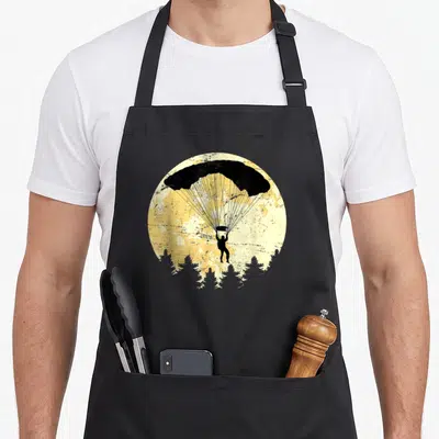 Tablier de cuisine noir avec illustration d'un parachutiste sous une lune jaune au-dessus d'arbres sombres.