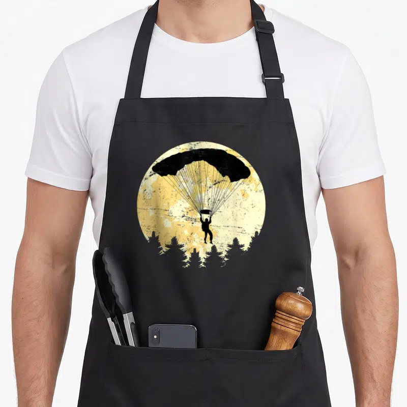 Tablier de cuisine noir avec illustration d'un parachutiste sous une lune jaune au-dessus d'arbres sombres.