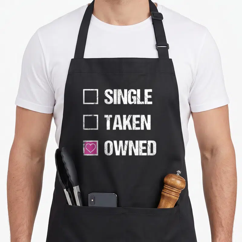 Tablier de cuisine noir avec motif textuel blanc et une case cochée rose contenant un cœur.