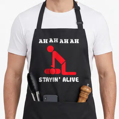 Tablier de cuisine noir avec une illustration rouge montrant un personnage effectuant un massage cardiaque sous les textes blancs Ah Ah Ah et Stayin' Alive.