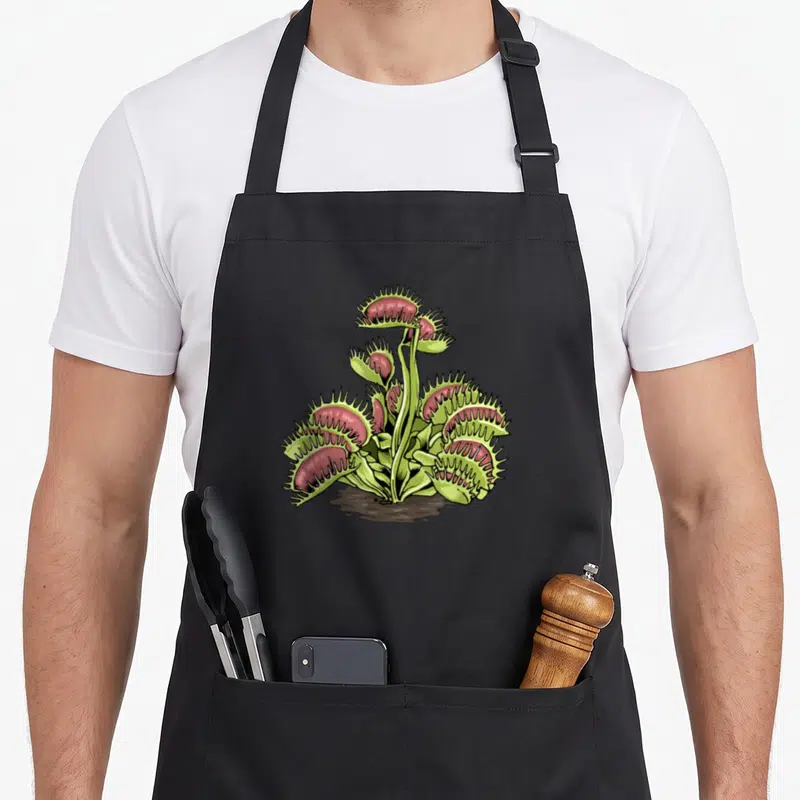 Tablier de cuisine noir avec une mouchette rose et verte aux pinces dentelées sortant du sol.