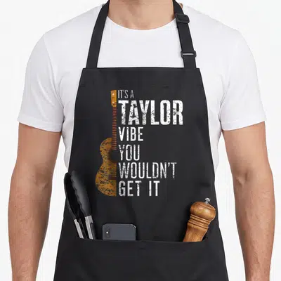 Tablier de cuisine noir avec une guitare acoustique orange et le texte blanc It's a Taylor ambiance you wouldn't get it.