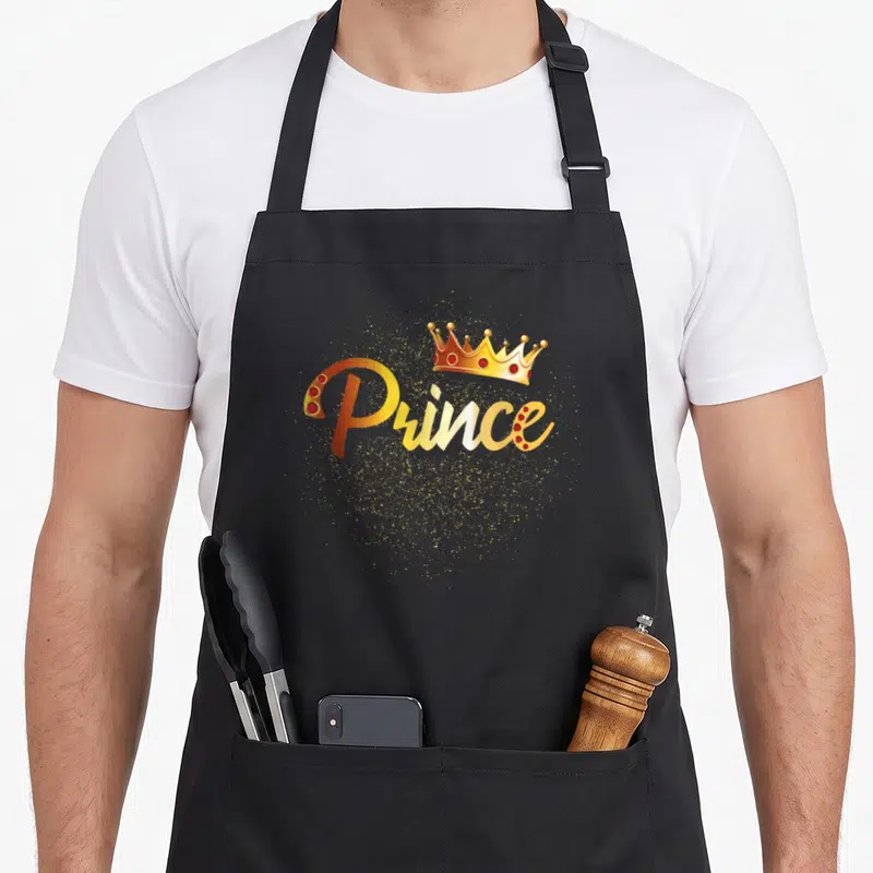 Tablier de cuisine noir avec inscription Prince dorée, couronne rouge et or, et éclats scintillants.