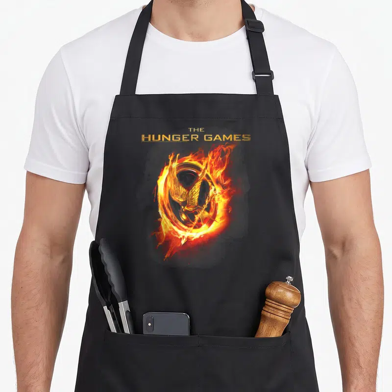 Tablier de cuisine noir avec un moqueur enflammé et le titre The Hunger Games en lettres dorées.