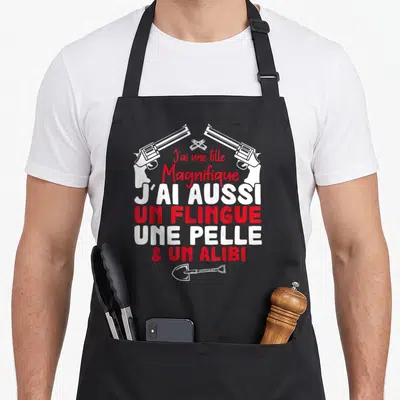 Tablier de cuisine noir avec inscription J'ai une fille magnifique et un alibi surmonté de deux revolvers croisés et d'une pelle en bas.