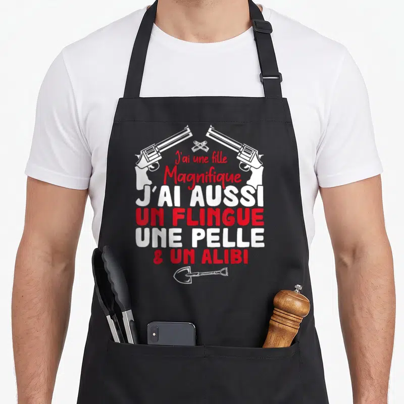 Tablier de cuisine noir avec inscription J'ai une fille magnifique et un alibi surmonté de deux revolvers croisés et d'une pelle en bas.